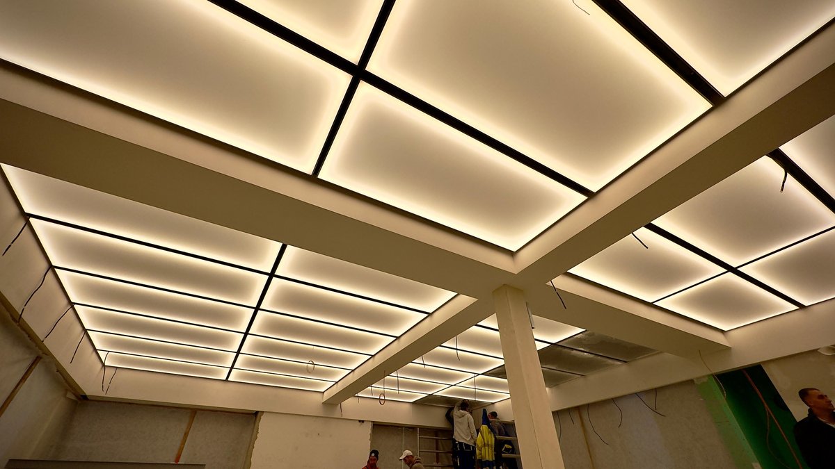 Moderní napínaný strop s integrovaným LED osvětlením v geometrickém designu s černými nosníky a rovnoměrně rozsvíceným světlem.