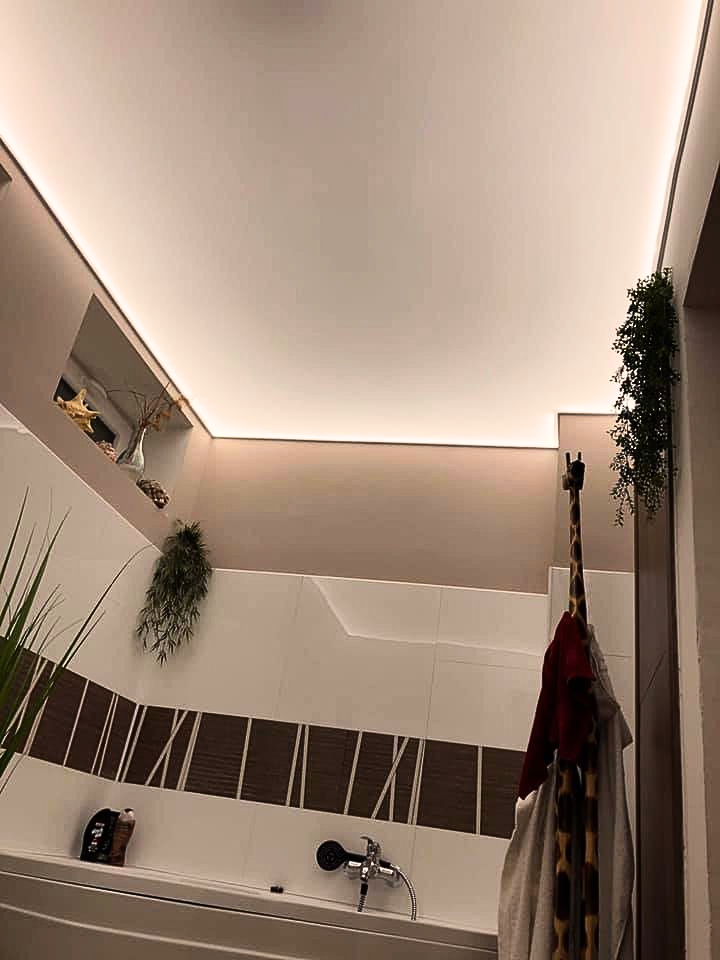 Koupelna s moderním napnutým stropem s integrovaným LED osvětlením a obkladem s dekorativním pruhem, doplněným zeleným rostlinami.