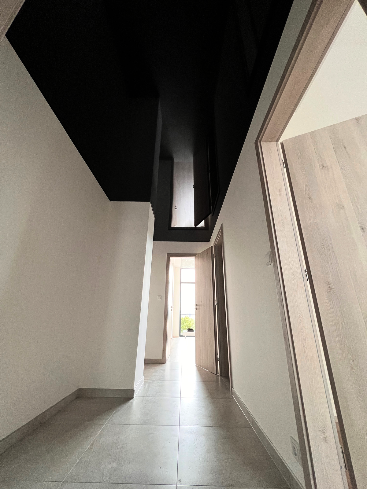 Dlouhá chodba s moderním černým napínaným stropem a integrovaným lineárním osvětlením, které vytváří dramatický kontrast s bílými stěnami a světlým podlahem.